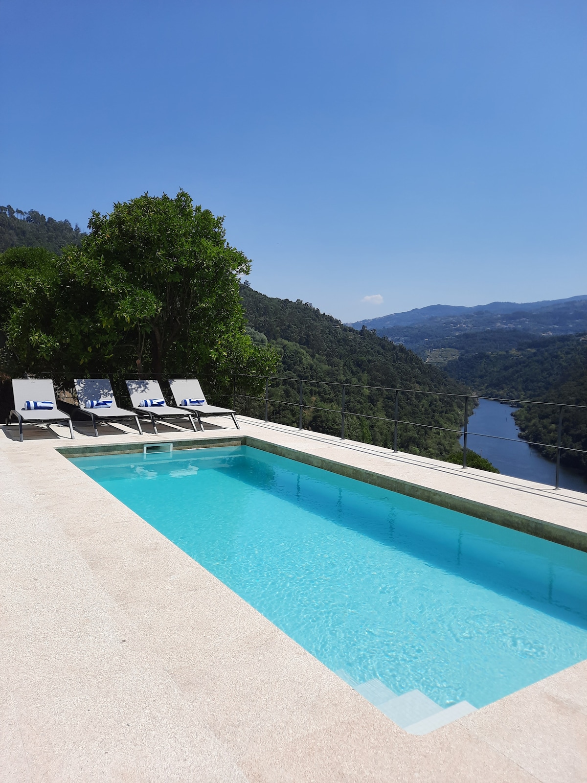 A private pool with clear turquoise water is set against a backdrop of rolling green hills and the Douro River. Four lounge chairs are positioned poolside, offering space for relaxation while enjoying the serene views of the surrounding landscape and distant mountains.