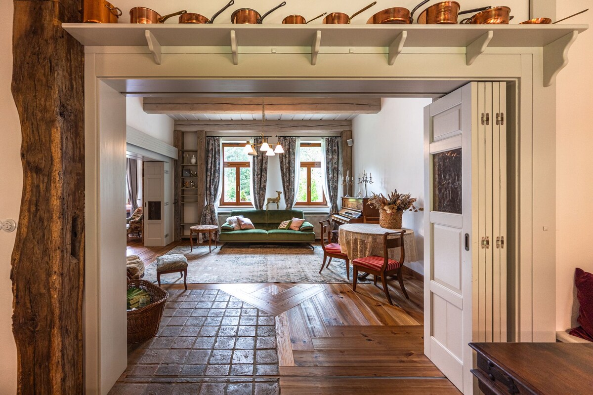 An open doorway reveals a spacious living area featuring warm wood accents and a blend of traditional and modern furnishings. Natural light floods through large windows, illuminating a cozy seating arrangement and a decorative table. Copper pots adorn the shelf above the doorway, adding character.