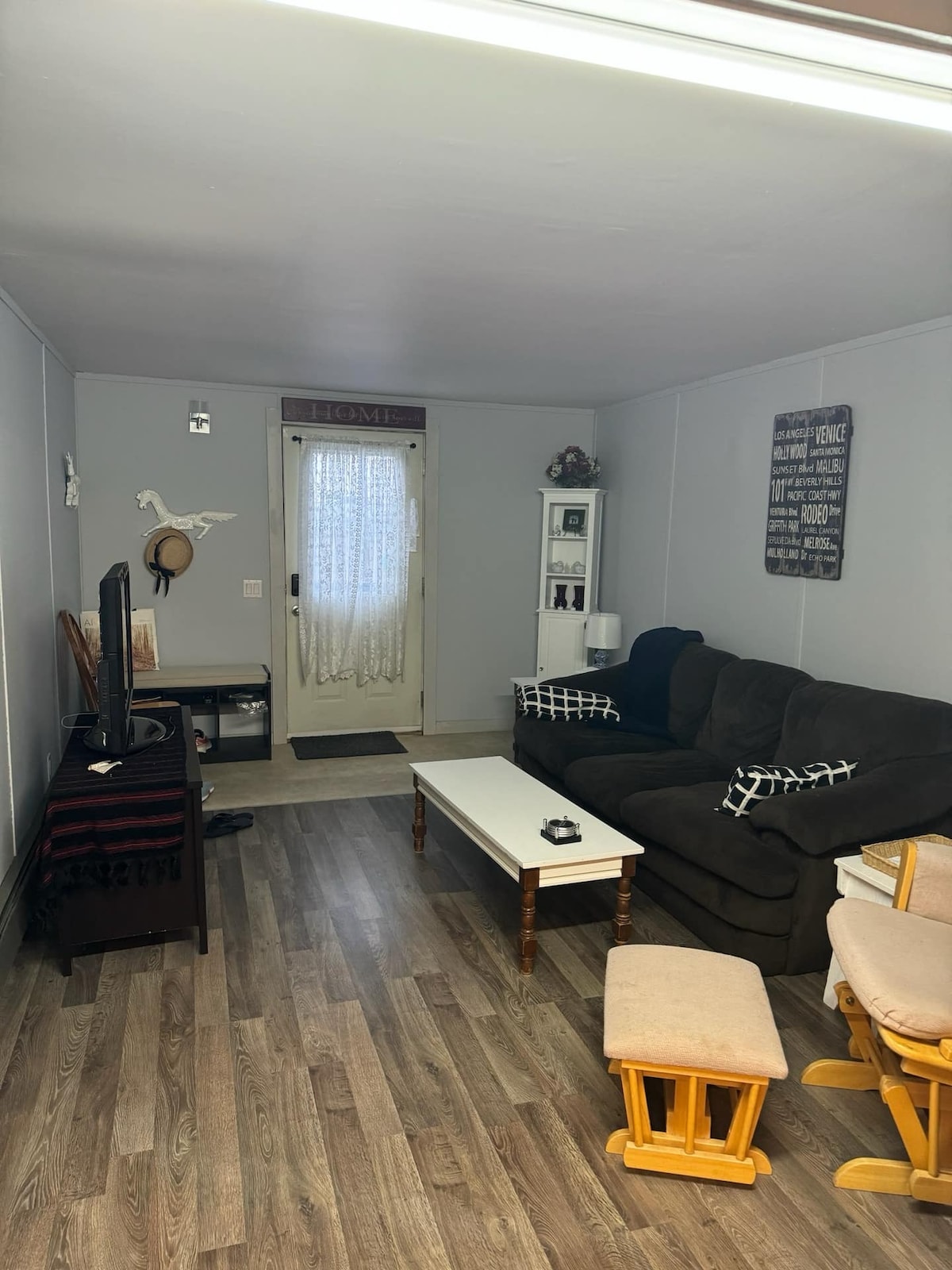 A living area features a dark sofa and a wooden coffee table, complemented by a small entertainment setup. A door with a lace curtain allows natural light, while decorative items are displayed on a nearby shelf. Wooden flooring extends throughout the space.