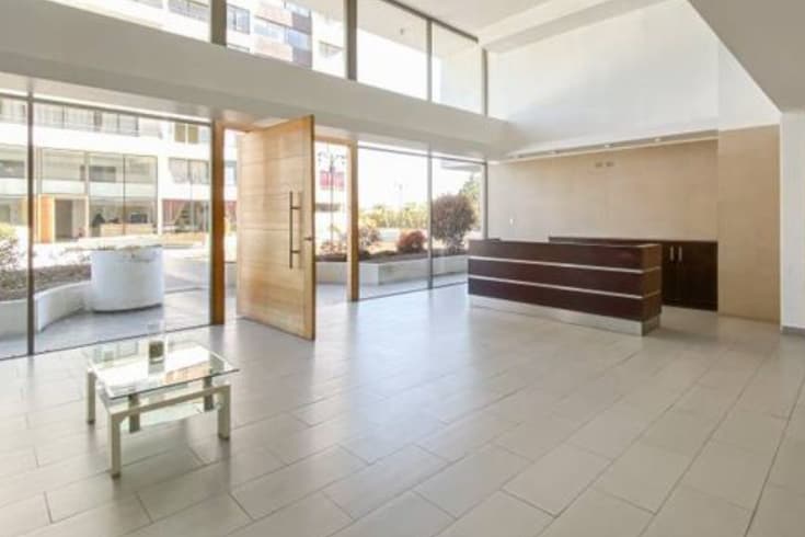 A spacious lobby area is highlighted by large glass windows, allowing ample natural light. A sleek reception desk stands against the wall, while minimalist furniture is arranged for casual seating. A small glass table in the center complements the modern design.