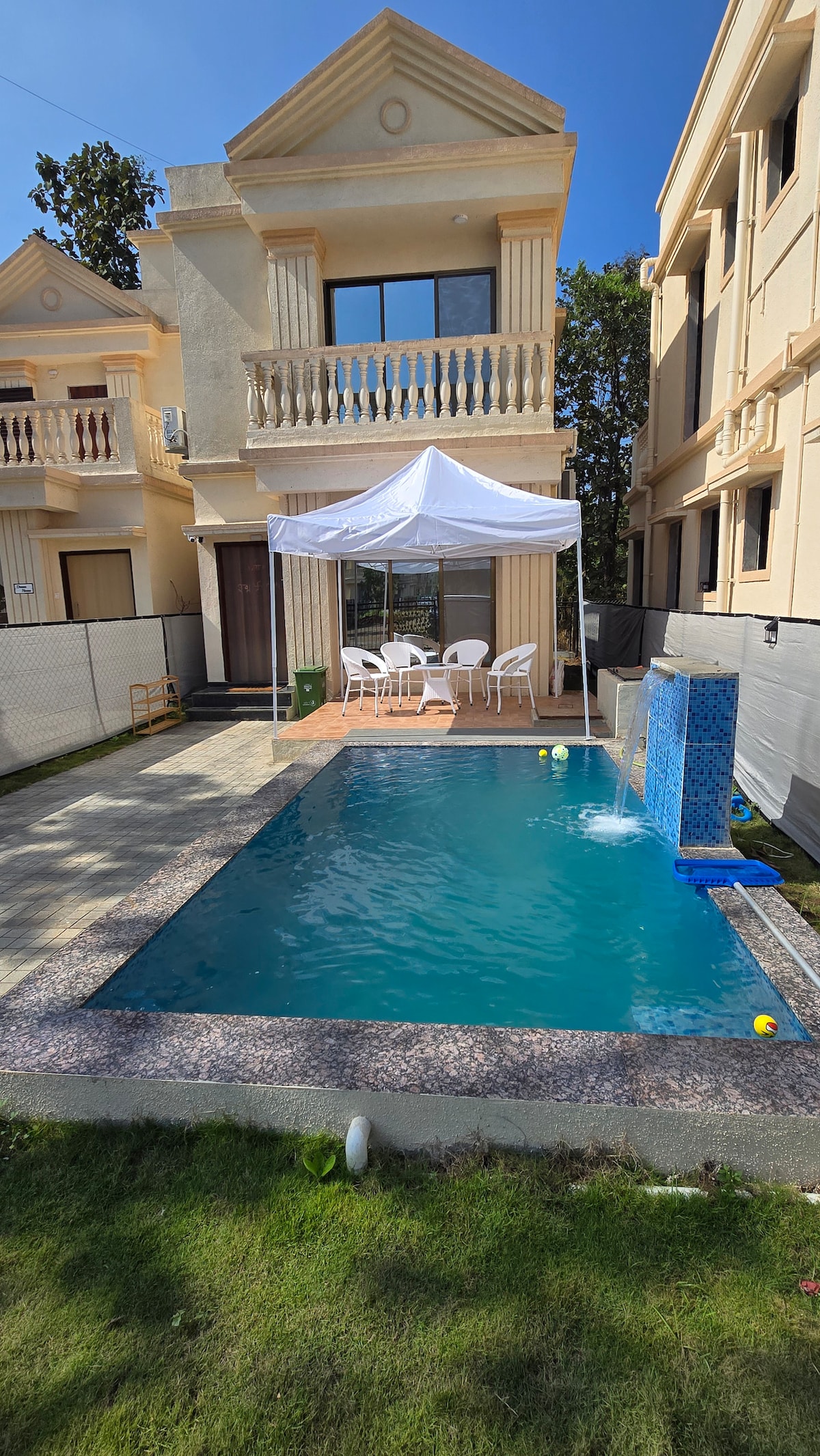 A private pool area is framed by a well-maintained lawn, featuring a tented seating arrangement. The pool, with a vibrant blue hue, is accompanied by colorful balls resting on the surface. Nearby, the villa displays a classic architectural design with a balcony above.