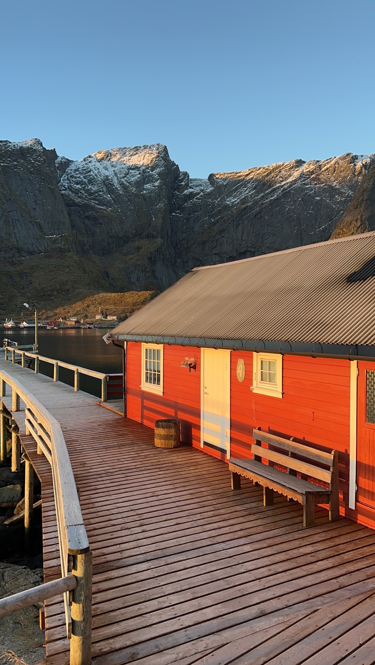 A wooden walkway extends towards a striking red building, showcasing a prominent entrance with a white door. The backdrop features majestic mountains, partially capped with snow, illuminated by the warm glow of sunlight.