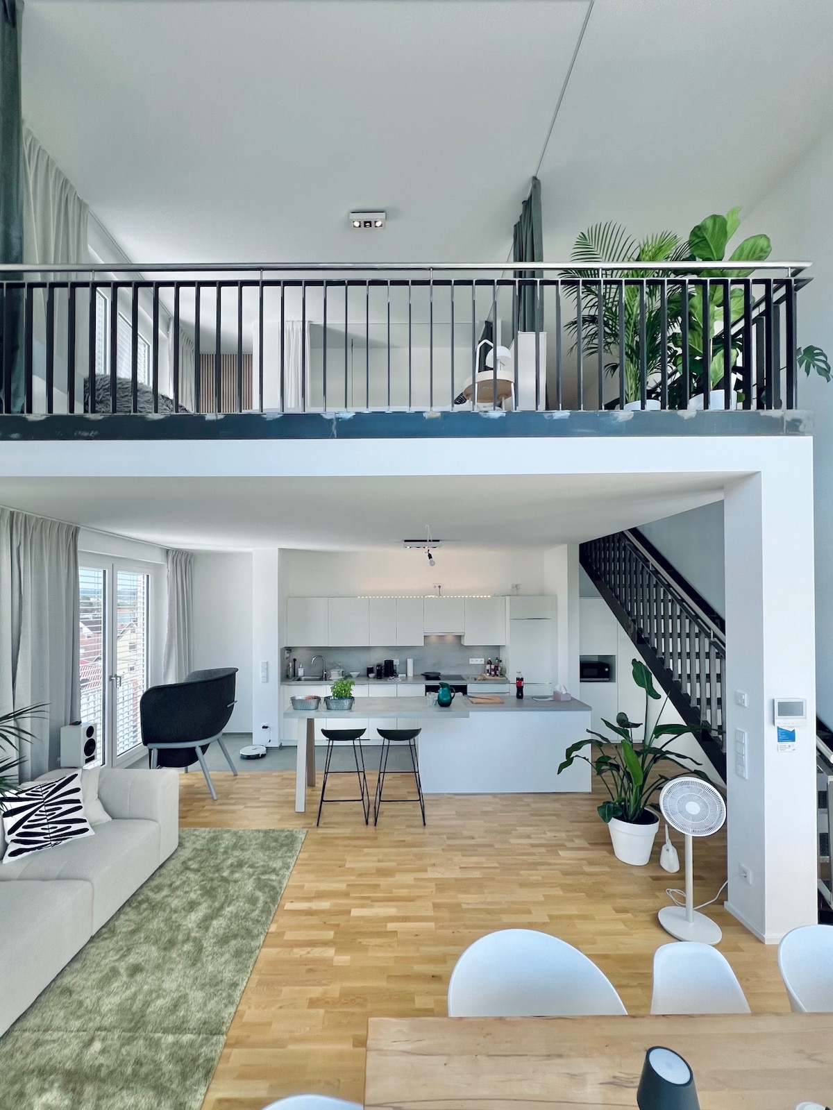 An open loft space is captured, showcasing a bright living area with light wood flooring and a comfortable sofa. A modern kitchen is visible in the background, complemented by high ceilings and greenery. A staircase leads to an upper level, enhancing the spacious design.