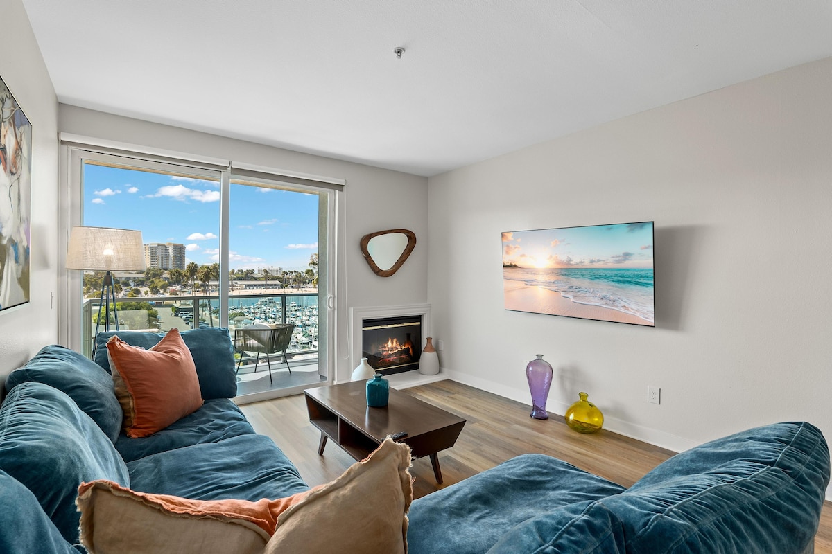 A spacious living area is presented with a plush sectional sofa in deep blue, accented by colorful pillows. A large-screen TV and a modern fireplace enhance the comfort. Floor-to-ceiling glass sliders open to a balcony, offering views of the marina and beach.