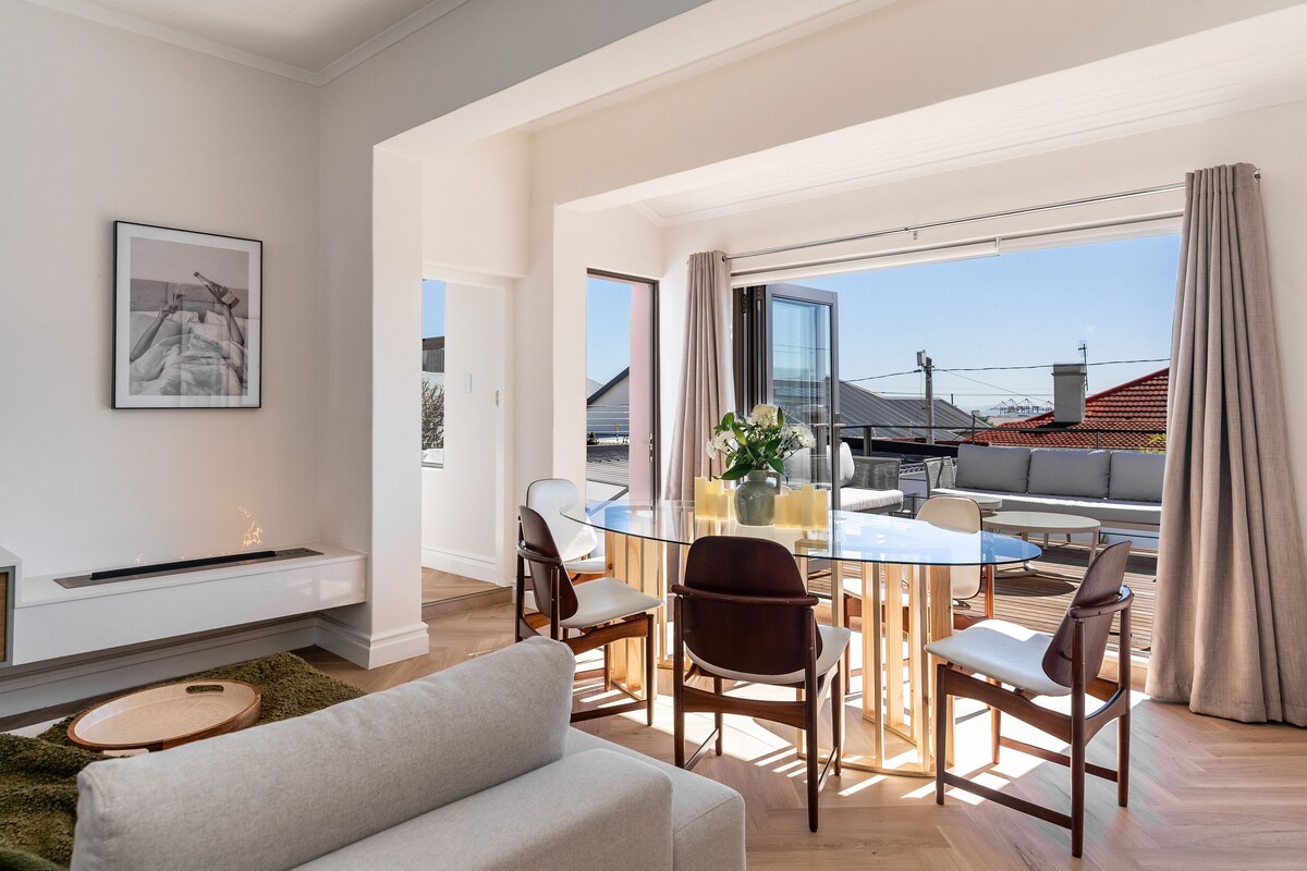An open-plan living and dining area is illuminated by natural light flowing through large glass doors. A circular glass dining table is accompanied by wooden chairs, while a comfortable sofa is positioned nearby. Warm tones and textures create a relaxed ambiance.