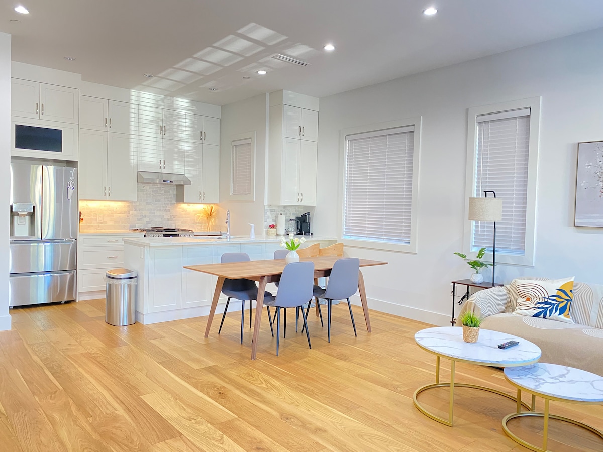 An open-plan living and dining area is revealed, showcasing a modern kitchen equipped with stainless steel appliances. A wooden dining table is surrounded by four gray chairs, while a cozy seating area features a light couch with colorful accents. Natural light filters through the windows, enhancing the warm wooden flooring.