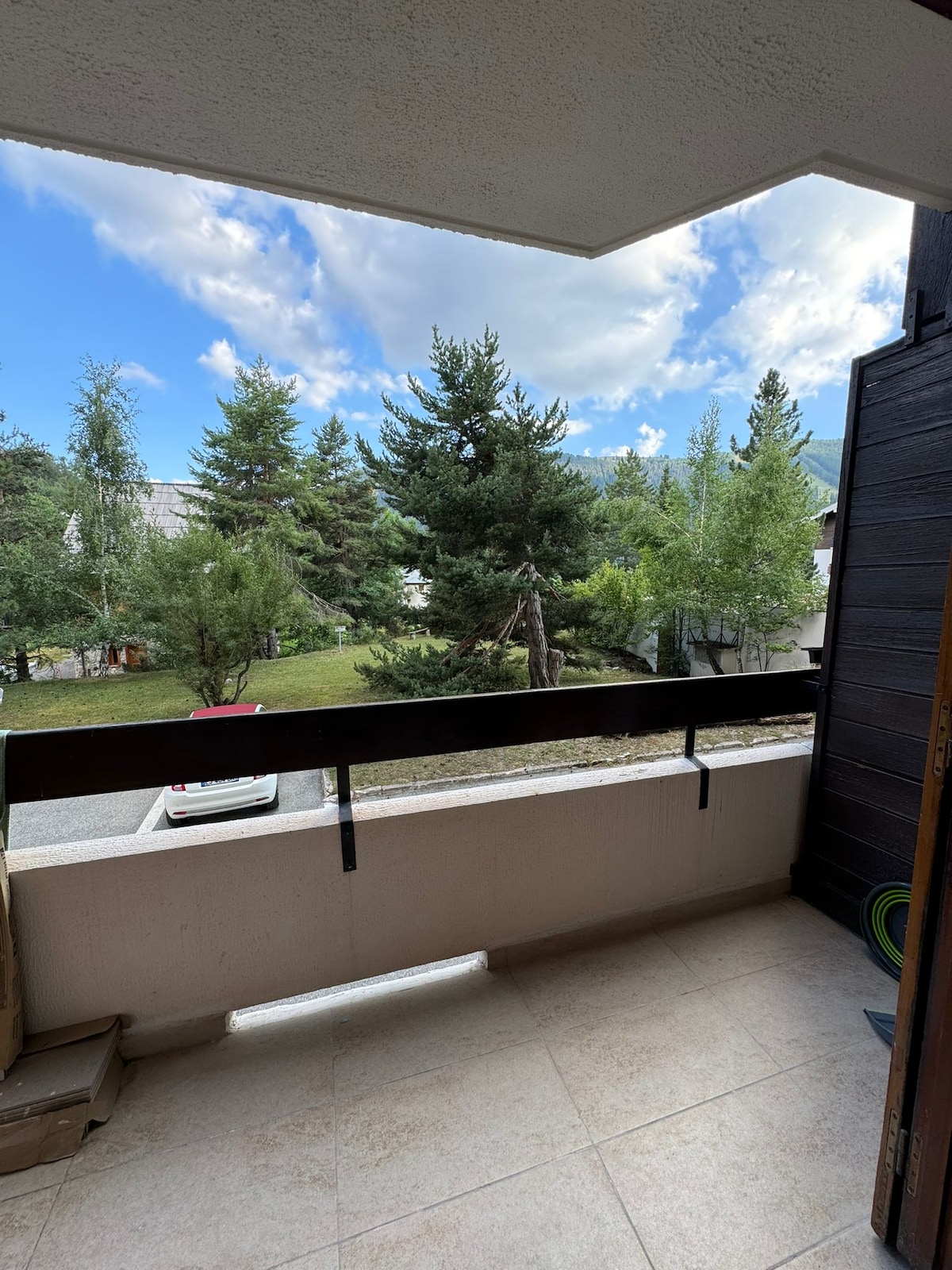 A balcony offers a view of lush greenery and tall trees, set against a backdrop of blue skies with clouds. A smooth tiled floor provides an inviting space, while a black railing ensures safety and security. The surroundings display a serene and natural environment.