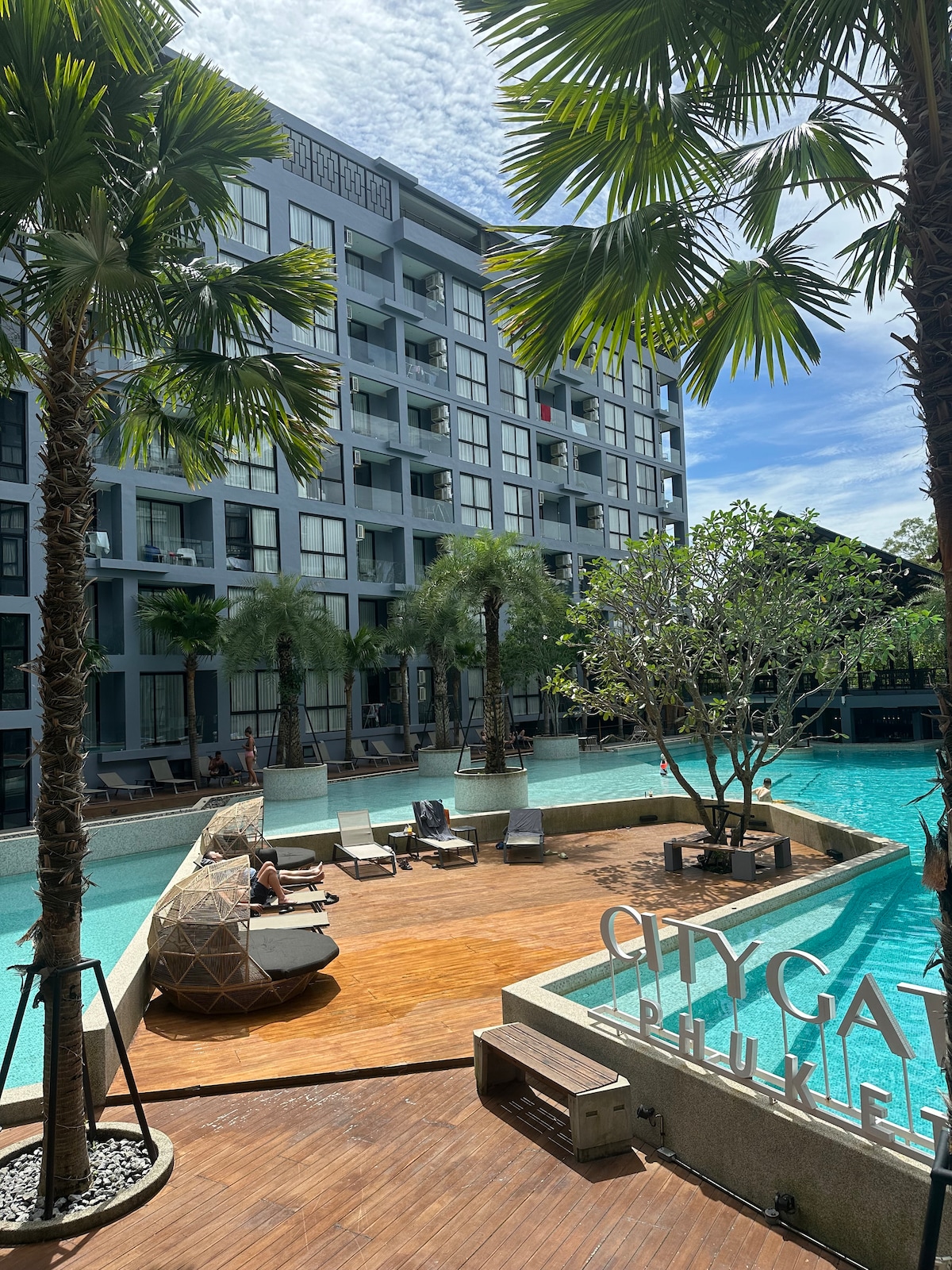 A serene pool area is surrounded by lush palm trees and modern architecture. Lounge chairs are positioned along the water's edge, and a wooden deck extends into the pool, enhancing the tranquil setting of the space.