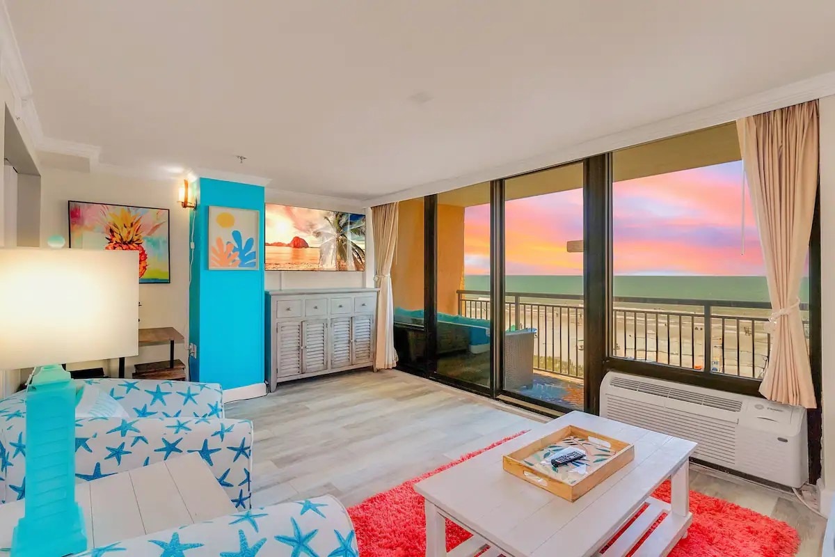 The living room features large floor-to-ceiling sliding glass doors offering expansive ocean views. A colorful accent wall is complemented by light-toned furniture, including a cozy couch and a modern coffee table. Soft lighting adds a warm touch to the inviting atmosphere.