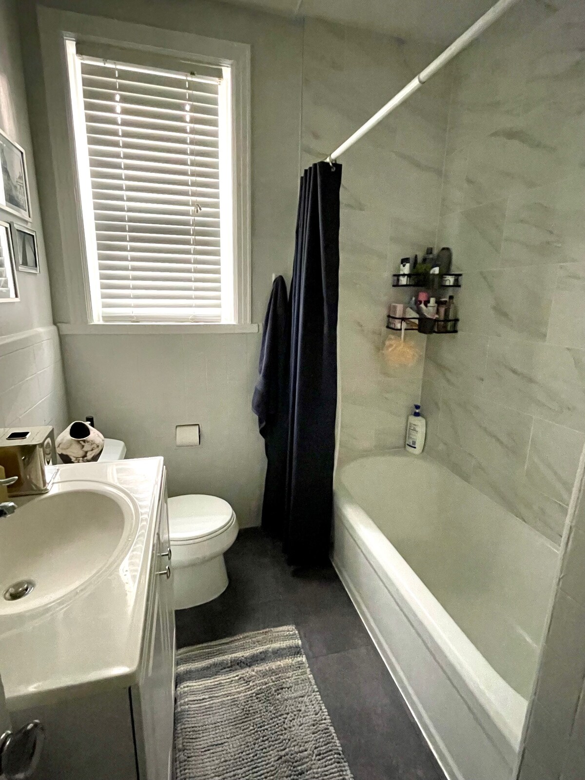 A recently renovated bathroom features a white bathtub with an attached shower curtain and modern toiletries arranged on a wall-mounted shelf. A sleek vanity with a basin is present, alongside a toilet. Natural light enters through a window with horizontal blinds above.