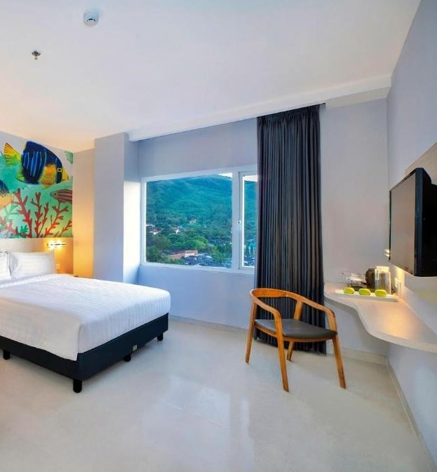 The room features a spacious bed against a backdrop of colorful wall art, capturing marine life. Natural light filters through a large window, which frames a view of lush hills. A wooden chair and a small table are positioned beside the bed.