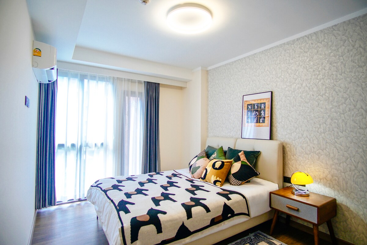 A bright bedroom features a spacious bed adorned with patterned blankets and decorative pillows. Natural light filters through sheer curtains, illuminating the room's textured wall. A bedside table holds a yellow lamp, complementing the modern design of the space.