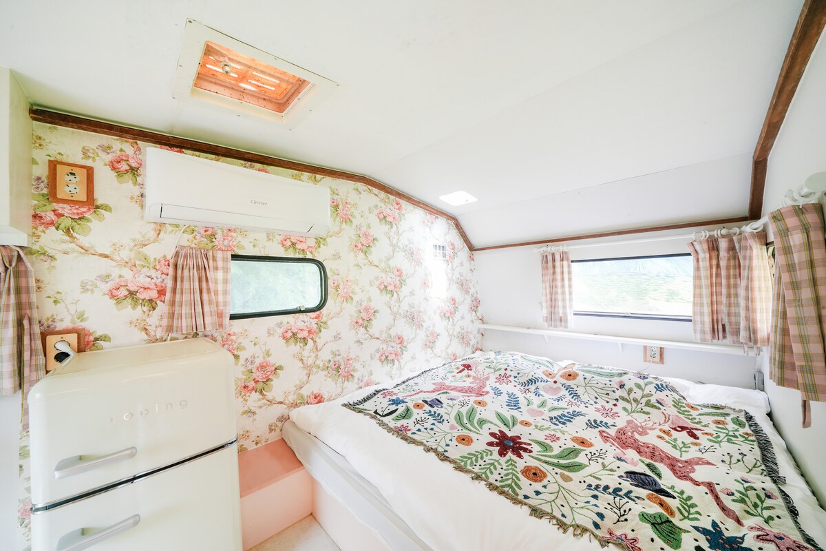 The interior of a vintage caravan features floral wallpaper and natural light streaming through two windows. A spacious bed is adorned with a colorful quilt, while a small refrigerator stands nearby. Air conditioning is provided for comfort, enhancing the serene atmosphere.