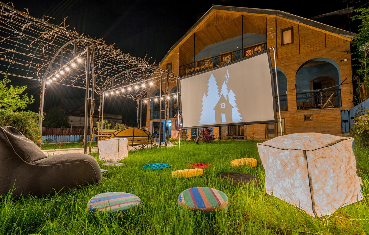 An outdoor movie screening area is set up on a grassy lawn, featuring a large screen draped between two structures. Colorful poufs and bean bags are arranged for seating, illuminated by string lights overhead. The surrounding building is constructed from warm-toned bricks.