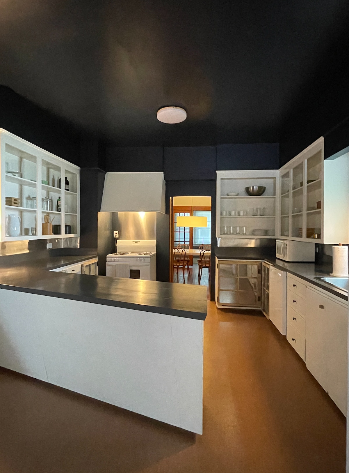 The kitchen features sleek metal countertops and white cabinetry with glass-front doors. Open shelving displays dishes, while a modern stove is positioned nearby. Soft light illuminates the space from a ceiling fixture, creating a bright and functional area with a view into the adjoining dining area.