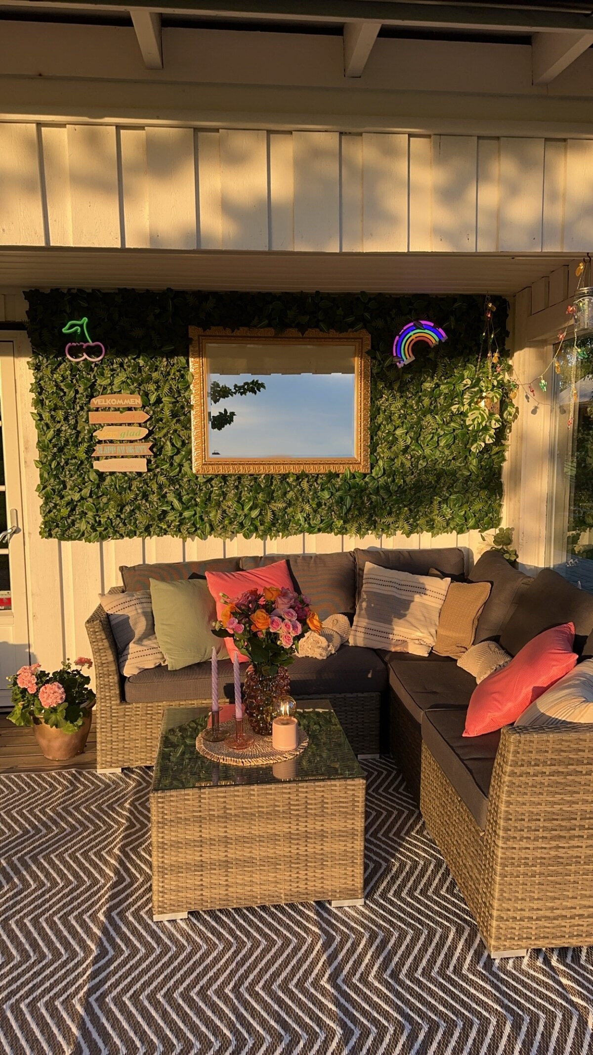 A cozy seating area features a rattan-style sofa adorned with various cushions in soft colors. A glass coffee table sits in front, complemented by a vase of flowers. A lush green wall with decorative accents includes a mirror and playful light fixtures, illuminated by warm evening sunlight.