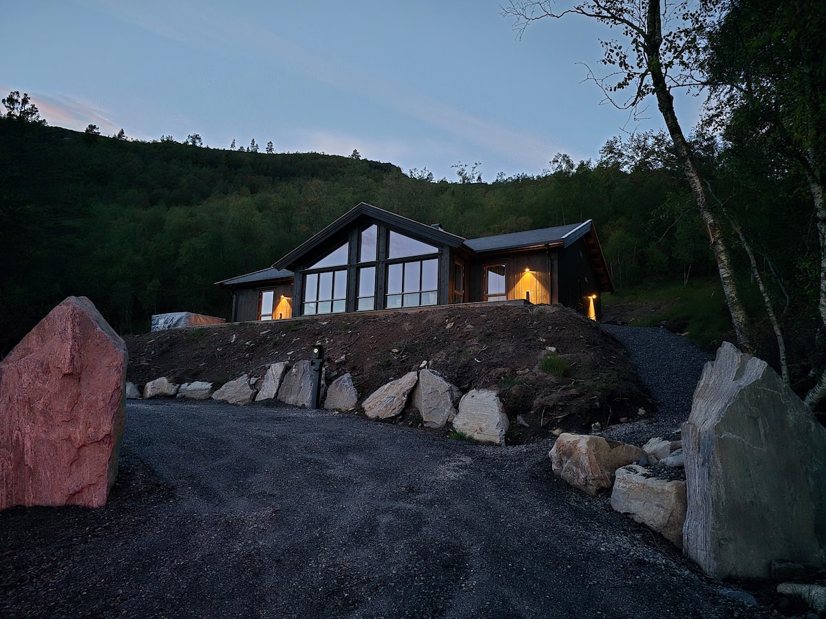A modern cabin is showcased against a twilight sky, with large glass windows reflecting the natural surroundings. Two warm lights glow from the entryway, inviting guests. The property is framed by large landscaping rocks, adding to the serene atmosphere.