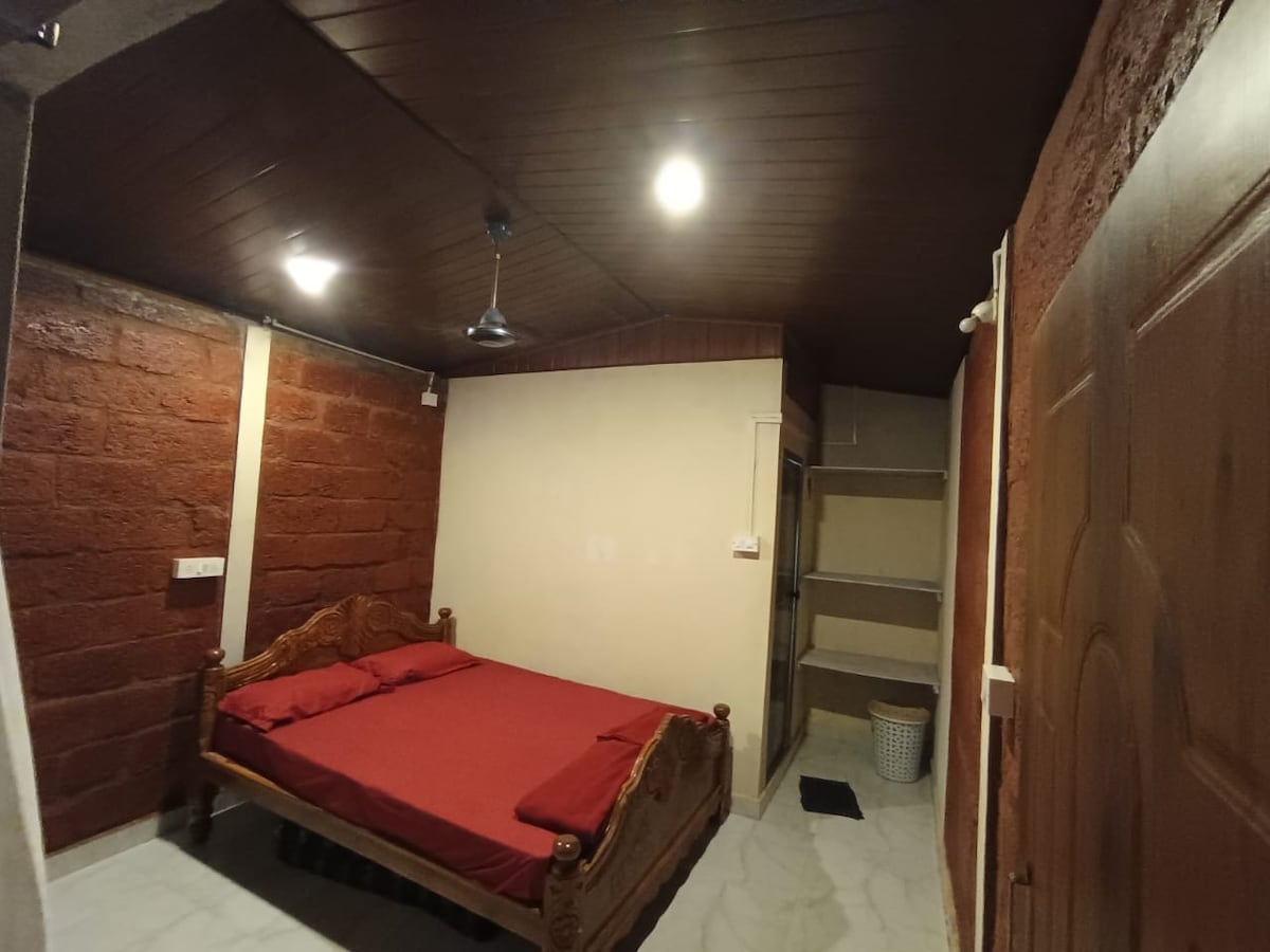 A compact bedroom features a wooden bed with a red bedspread, positioned against a textured stone wall. The ceiling is lined with dark wood, and bright ceiling lights illuminate the space. A small shelving unit is visible beside the bed, offering additional storage.