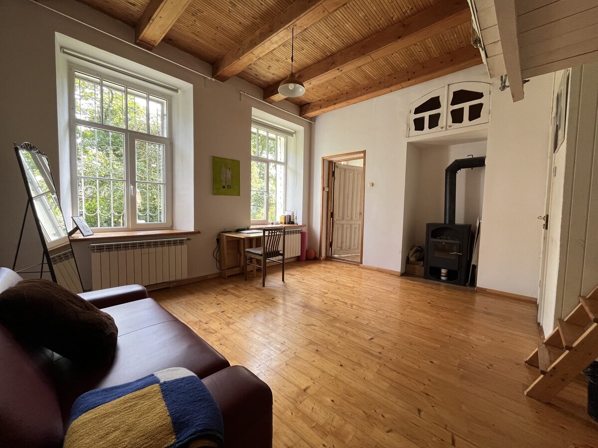 A spacious living area features high ceilings and large windows allowing natural light to fill the room. A wooden floor enhances the open atmosphere, complemented by a cozy sofa and a simple dining table set near a fireplace. A staircase leads to additional areas.
