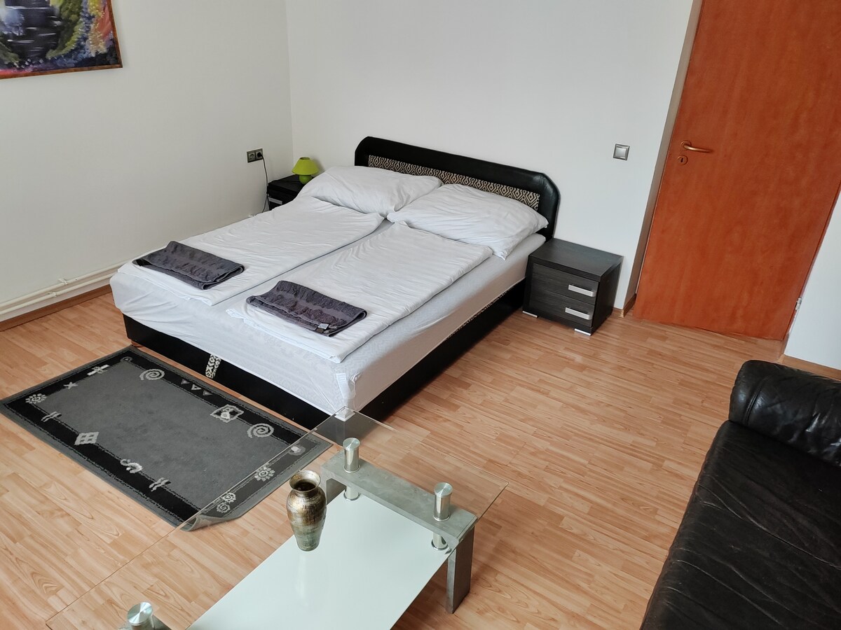 A spacious bedroom features a large bed with light linens. Two towels are neatly arranged at the foot of the bed. A glass coffee table is positioned in front of a dark sofa, and a small nightstand sits beside the bed. Natural light enters through the door.