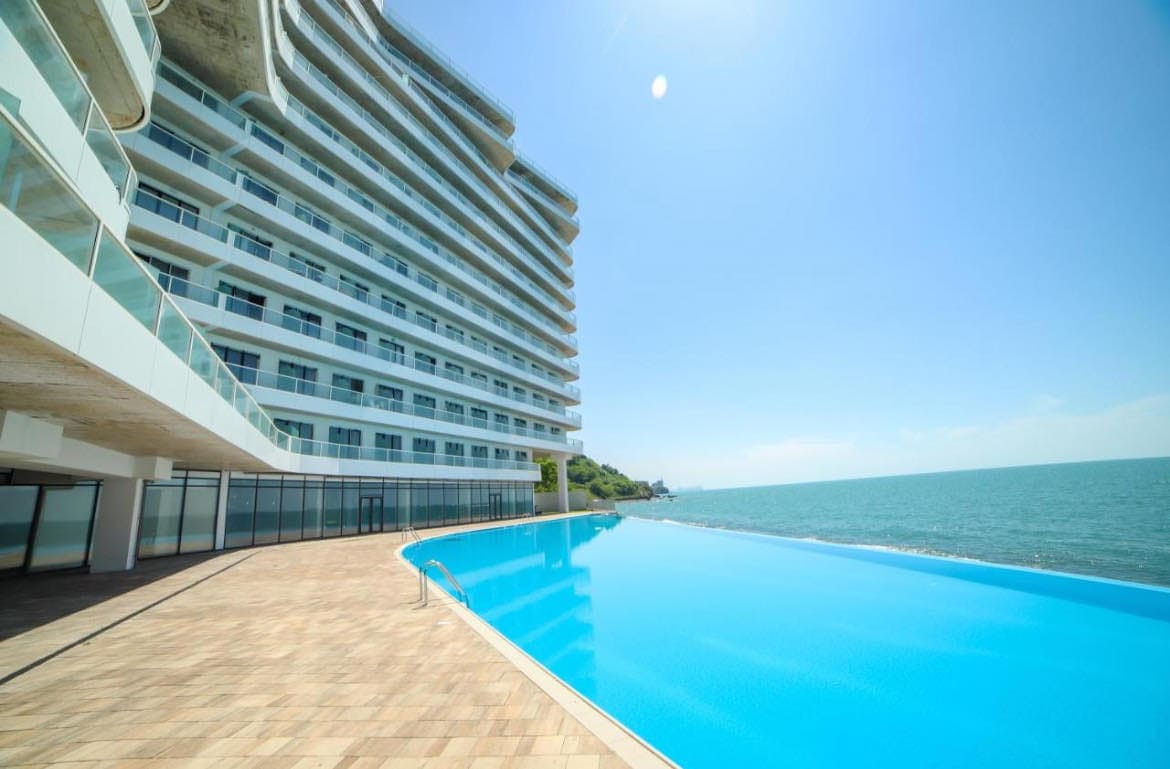 A large infinity pool stretches alongside a modern seaside building, providing a seamless view of the ocean. Sunlight reflects off the water's surface, creating a calm and serene environment. The spacious deck features paving stones and glass railings, enhancing the unobstructed coastal scenery.