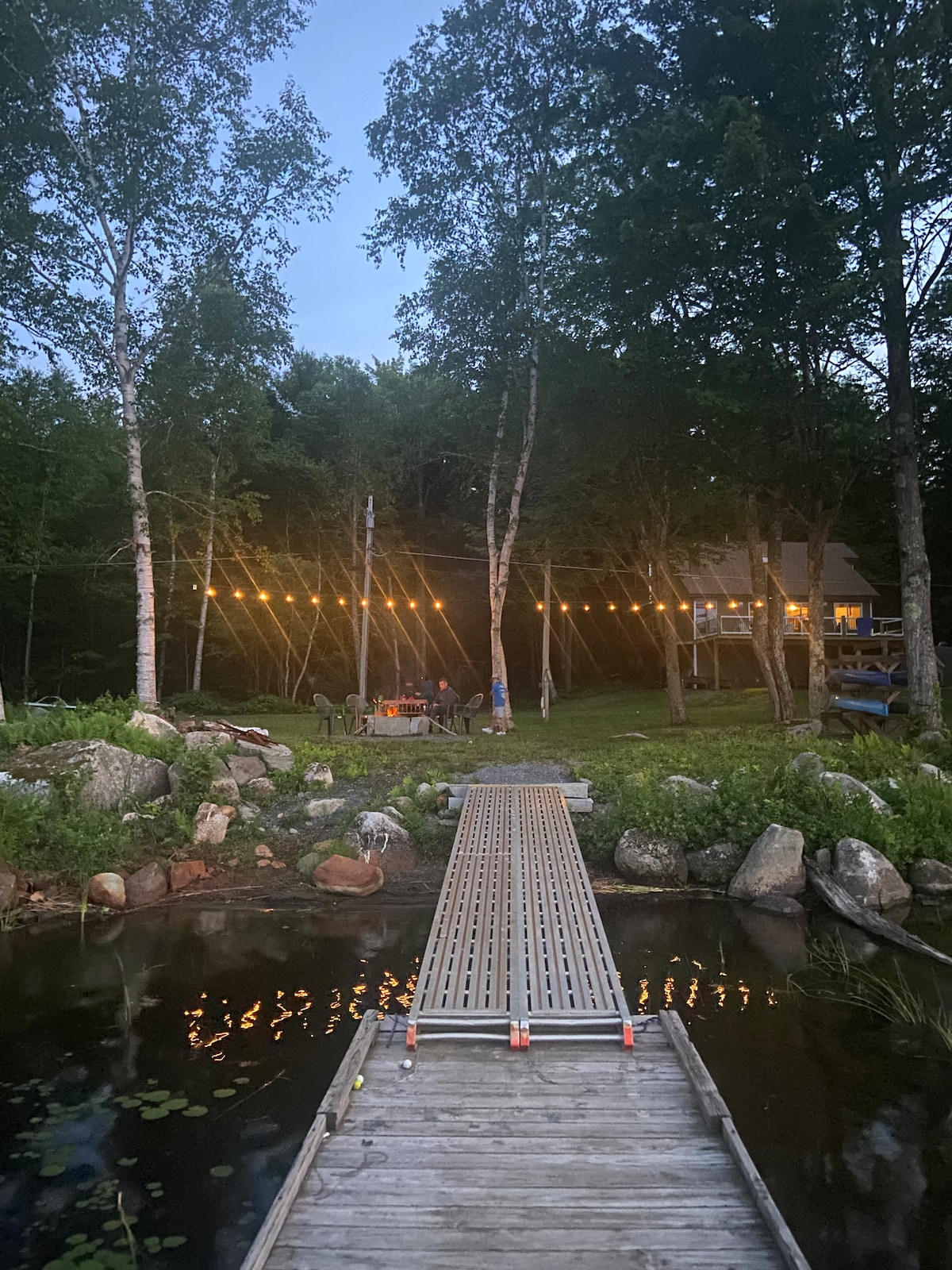 A wooden dock extends over calm waters, leading to a lush backyard illuminated by warm string lights. Tall trees and rocks accent the scene, while a gathering area with seating is visible in the distance, suggesting a space for socializing and relaxation.