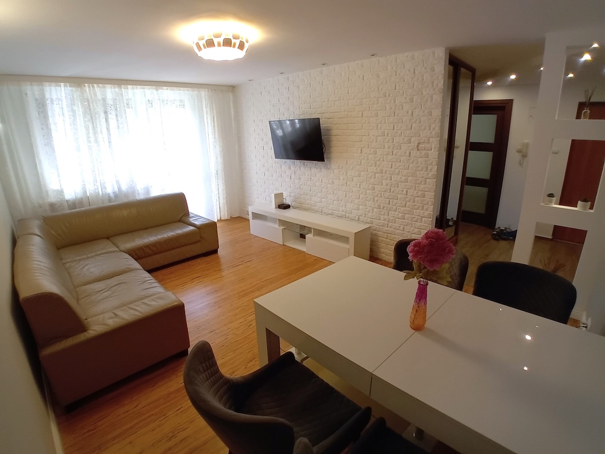 A spacious living area features a large sectional sofa and a modern coffee table. A wall-mounted television is positioned above a white media console. Natural light filters in through sheer curtains, illuminating the warm wooden floor.