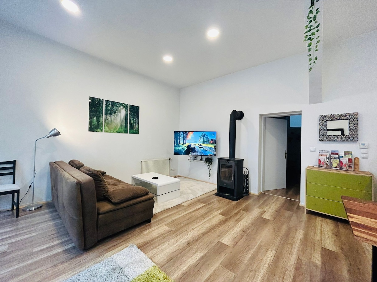 A modern living area is presented with a spacious brown sofa and a white coffee table. A large television is mounted on the wall, alongside two nature-themed artworks. A wood-burning stove adds warmth to the space, complemented by wooden flooring and green accents.