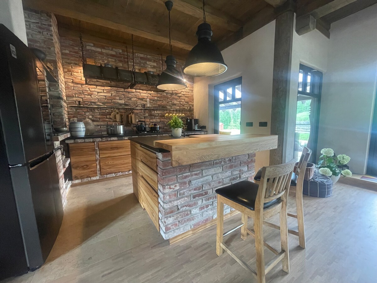A rustic kitchen space features a large wooden island with a brick facade and bar seating. The area includes modern appliances and shelves with kitchenware, illuminated by pendant lights. Large windows allow natural light to fill the room, enhancing the inviting atmosphere.