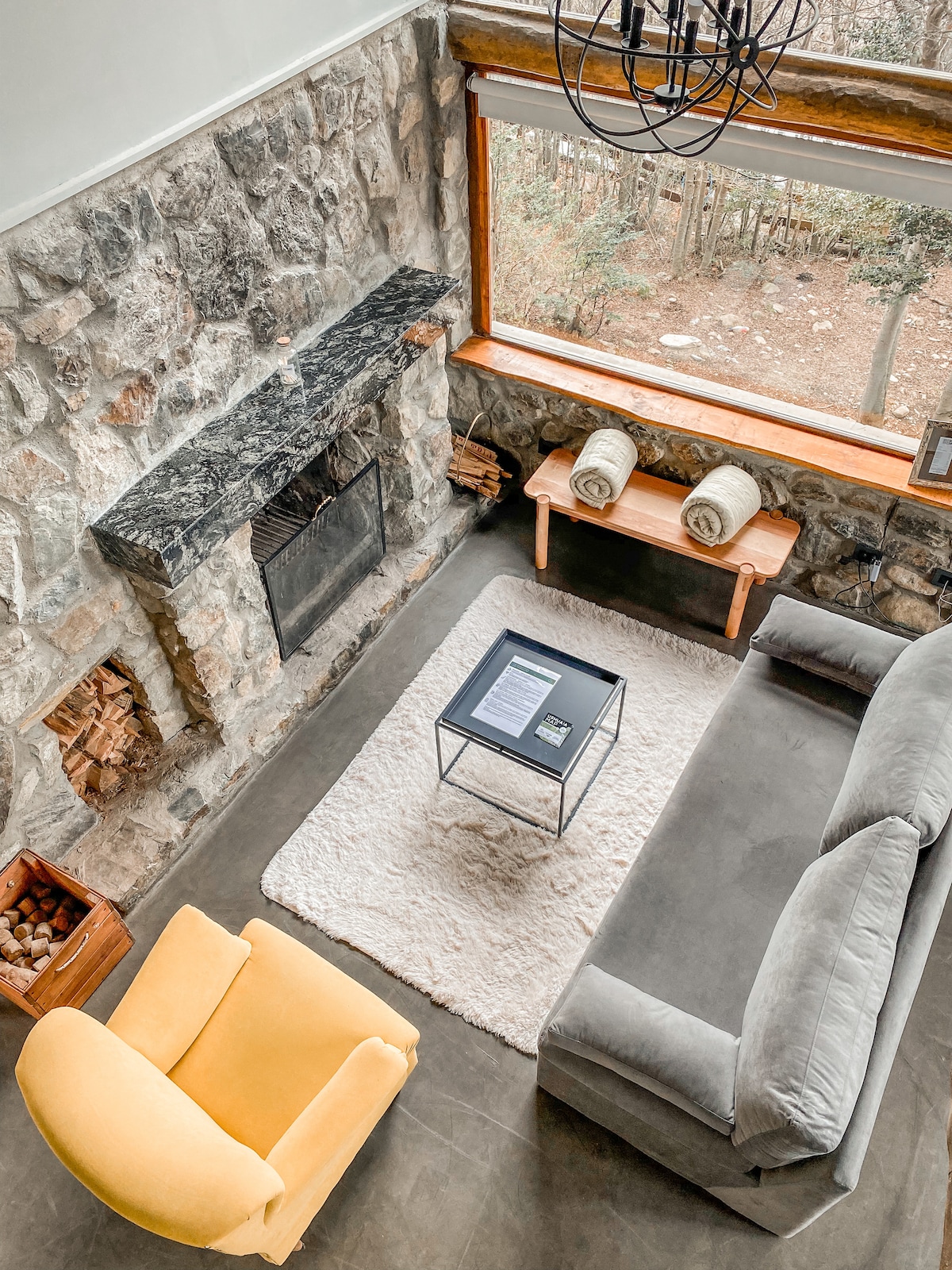 A living area features a gray sofa positioned next to a stone fireplace. A wooden table holds folded blankets, and a small black coffee table rests on a light rug. Large windows provide views of the surrounding trees, enhancing the serene atmosphere.