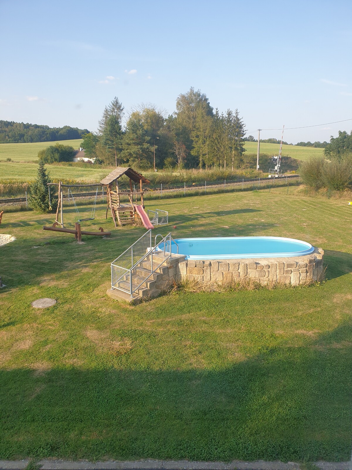 An outdoor area features a small swimming pool surrounded by natural grass. Nearby, a playground structure with a slide is visible, providing space for leisure activities. Trees line the background, enhancing the tranquil setting.