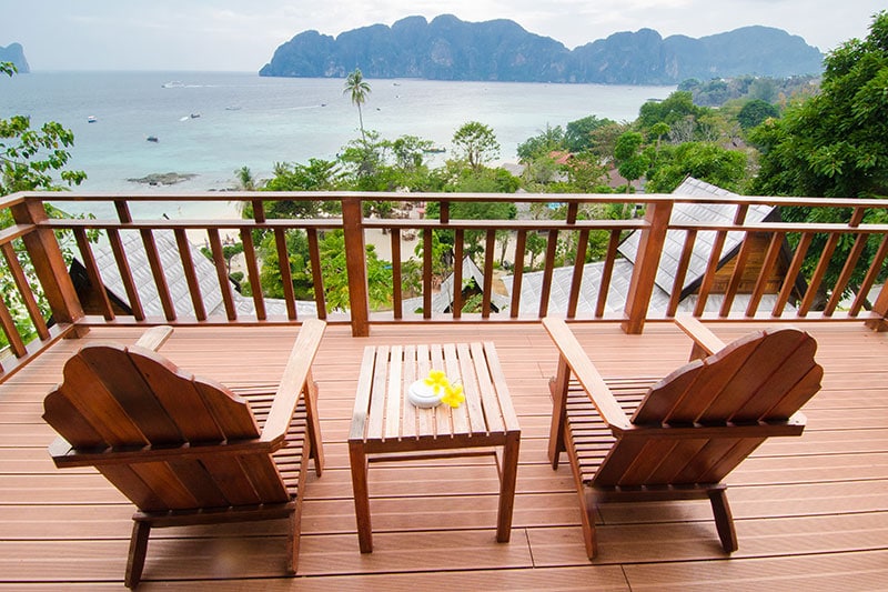 A wooden balcony overlooks the ocean and lush greenery, with two chairs placed around a small table. A plate with a flower rests on the table, while the distant coastline and hills provide a scenic backdrop.