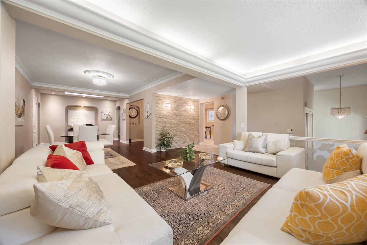 A spacious living area features a light-toned sectional sofa accented with colorful cushions and a decorative area rug. A glass coffee table sits at the center, while indirect lighting highlights the ceiling. An open dining space with a modern design is visible in the background.