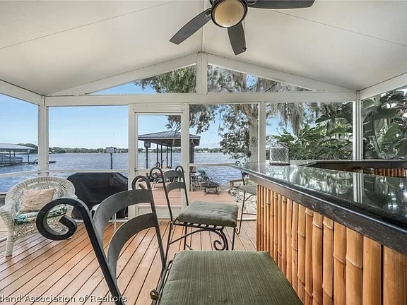 An inviting outdoor deck is showcased, featuring a bar area with black granite countertops and green cushioned stools. The view overlooks the tranquil waters of Lake Henry, framed by lush greenery and trees, creating a serene environment for relaxation and socializing.