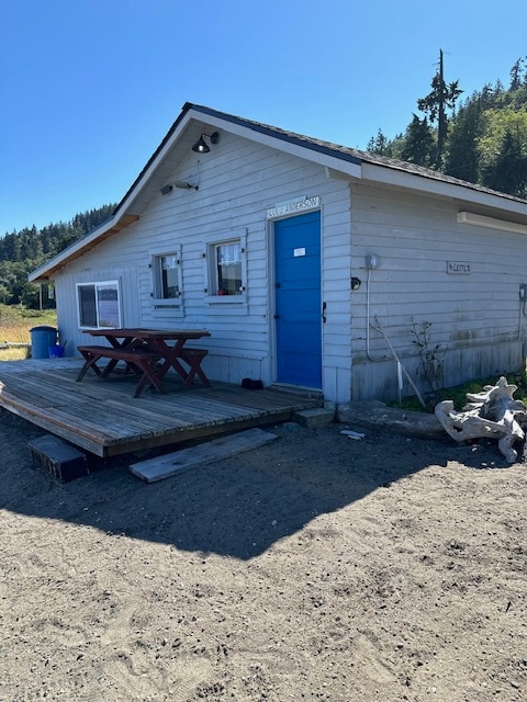 This waterfront cabin features a welcoming exterior with a bright blue door and multiple windows. A wooden deck includes a picnic table, positioned on a gravel pathway surrounded by nature. The roof slopes gently, and trees are visible in the background.