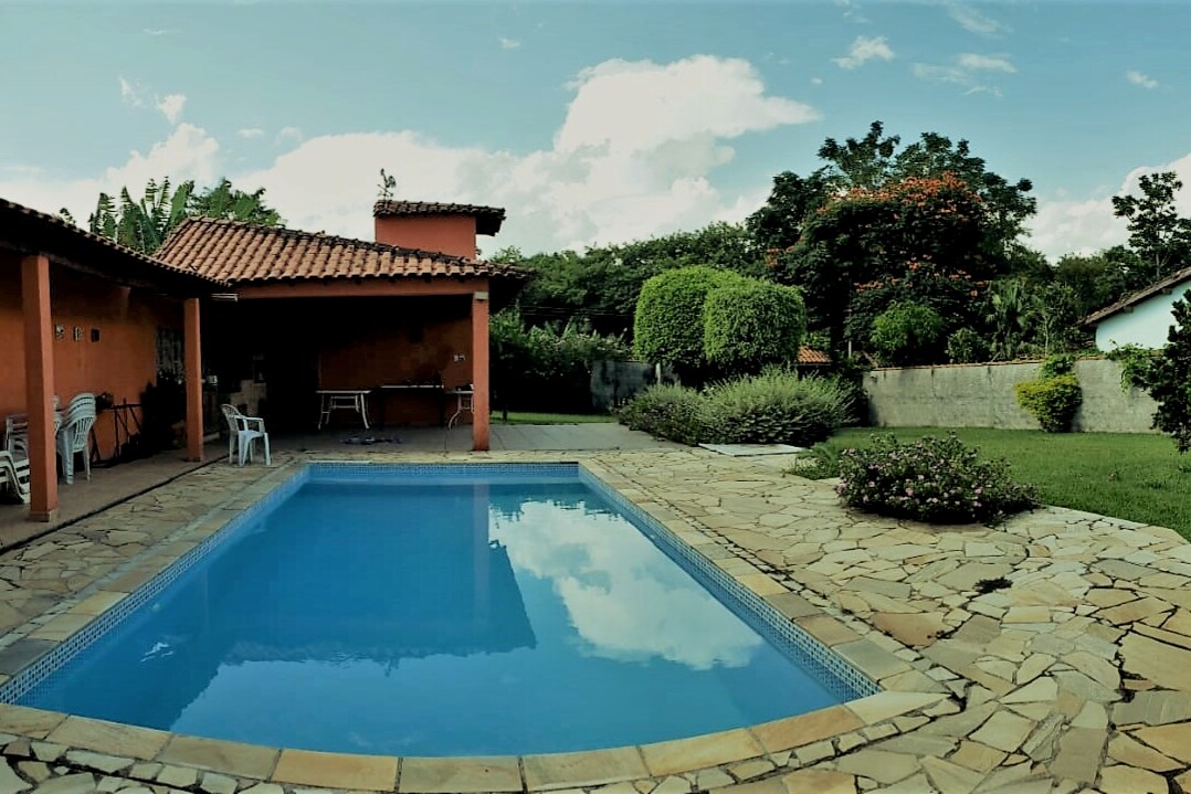 A calm swimming pool is framed by natural stone paving, reflecting the blue sky above. Lush greenery and flowering bushes surround the area, while a shaded patio with a terracotta roof offers a space for relaxation. White chairs provide additional seating nearby.