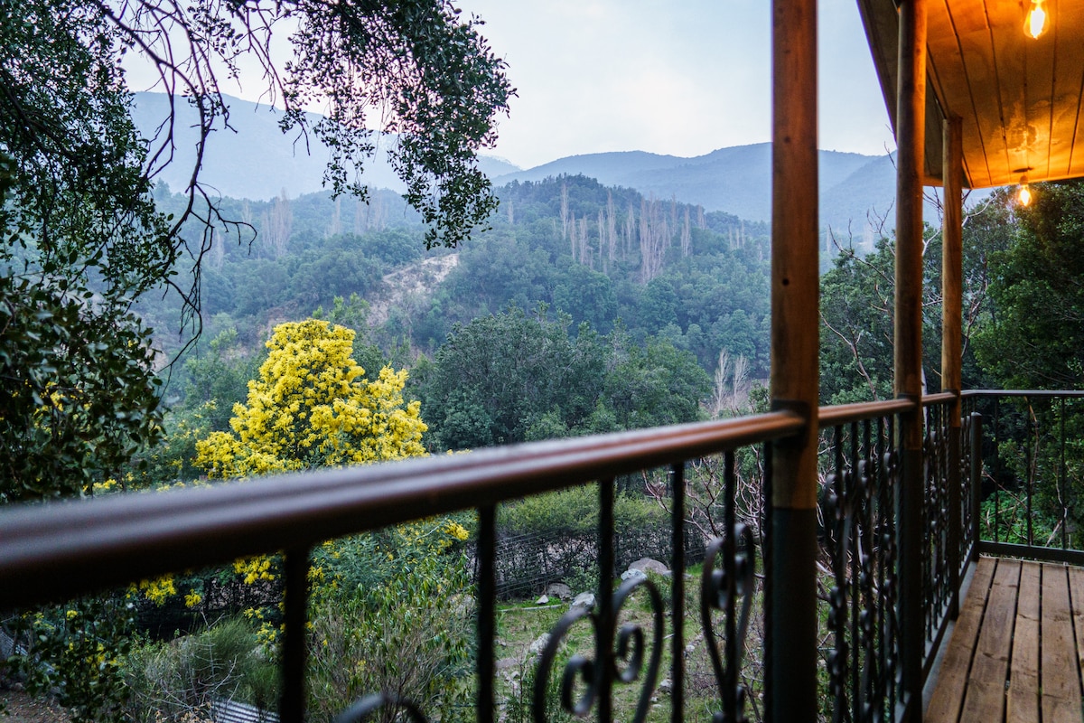 A balcony railing frames a serene view of the mountains, with lush greenery and trees visible. The landscape displays varying shades of green, with bright yellow foliage creating a focal point. Soft light suggests an early morning or late afternoon atmosphere.