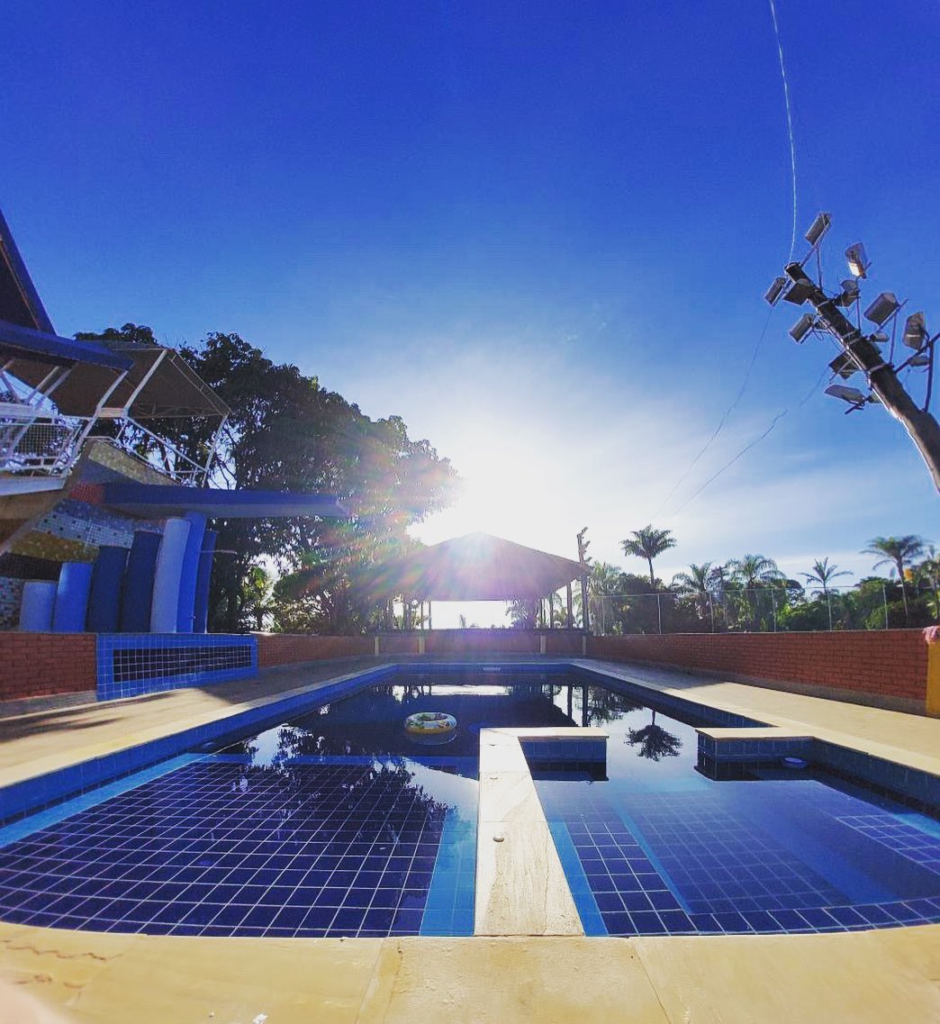 The pool area is surrounded by lush greenery and features a bright blue tiled pool reflecting the afternoon sun. A covered area is visible in the background, providing a shaded space, while palm trees frame the scene, creating a relaxing outdoor environment.
