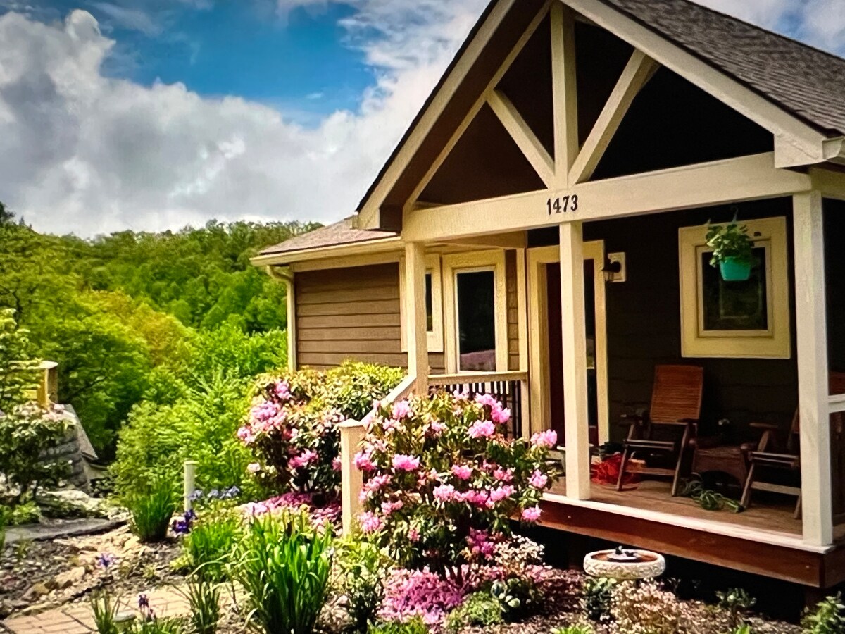 The entrance of the villa features a covered porch with two wooden rocking chairs, inviting relaxation. Lush greenery surrounds the porch, complemented by vibrant flowering plants in various colors, creating a cheerful and welcoming first impression.