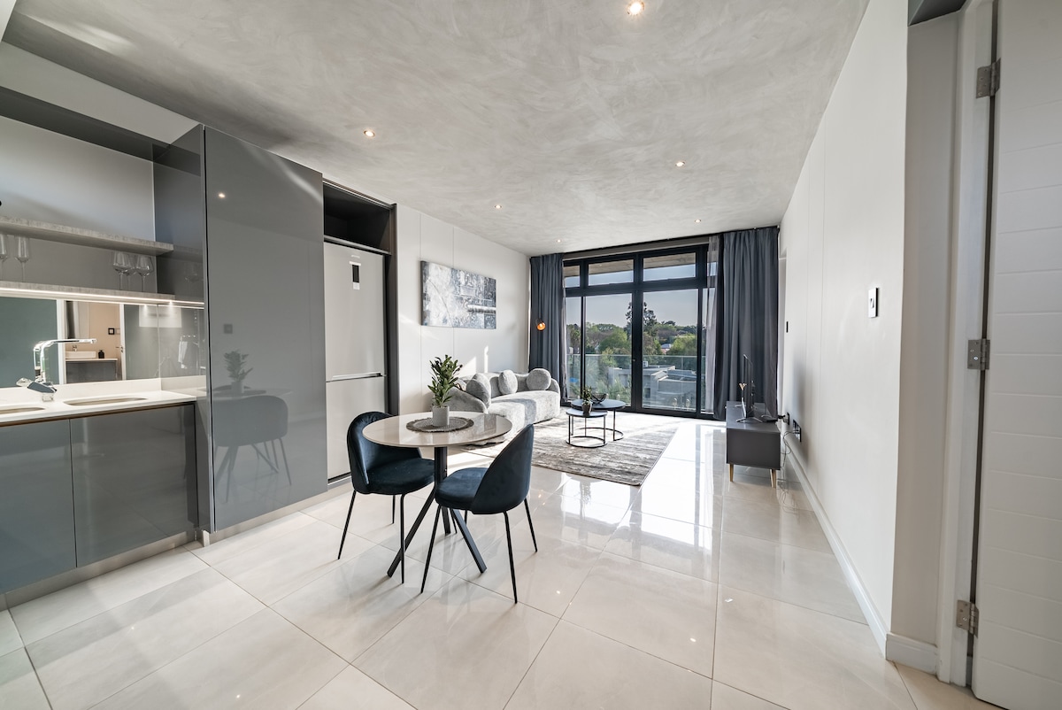 A modern living space is featured with white tiled flooring and ample natural light. The area includes a sleek kitchen with grey cabinetry, a dining table with four chairs, and a seating arrangement consisting of a sofa and armchair. Large glass doors open to a view of greenery.