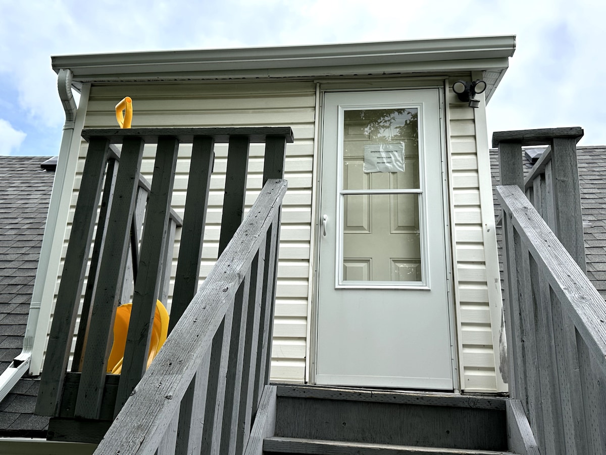 A private entrance is accessed via a set of wooden stairs, leading to a door framed with light-colored siding. A yellow object is visible beside the entrance, contributing a pop of color against the neutral tones.