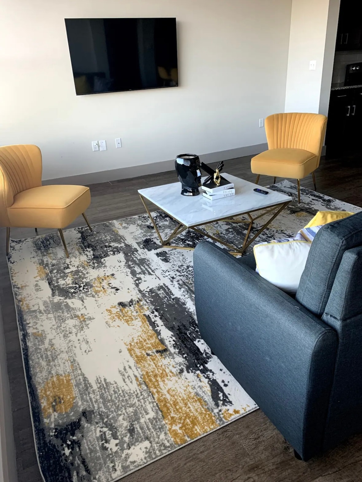 A modern living area is presented with two yellow armchairs and a dark gray sofa. A rectangular coffee table is centered on a patterned area rug, complemented by a flat-screen television mounted on the wall. Natural light fills the space, enhancing its open feel.