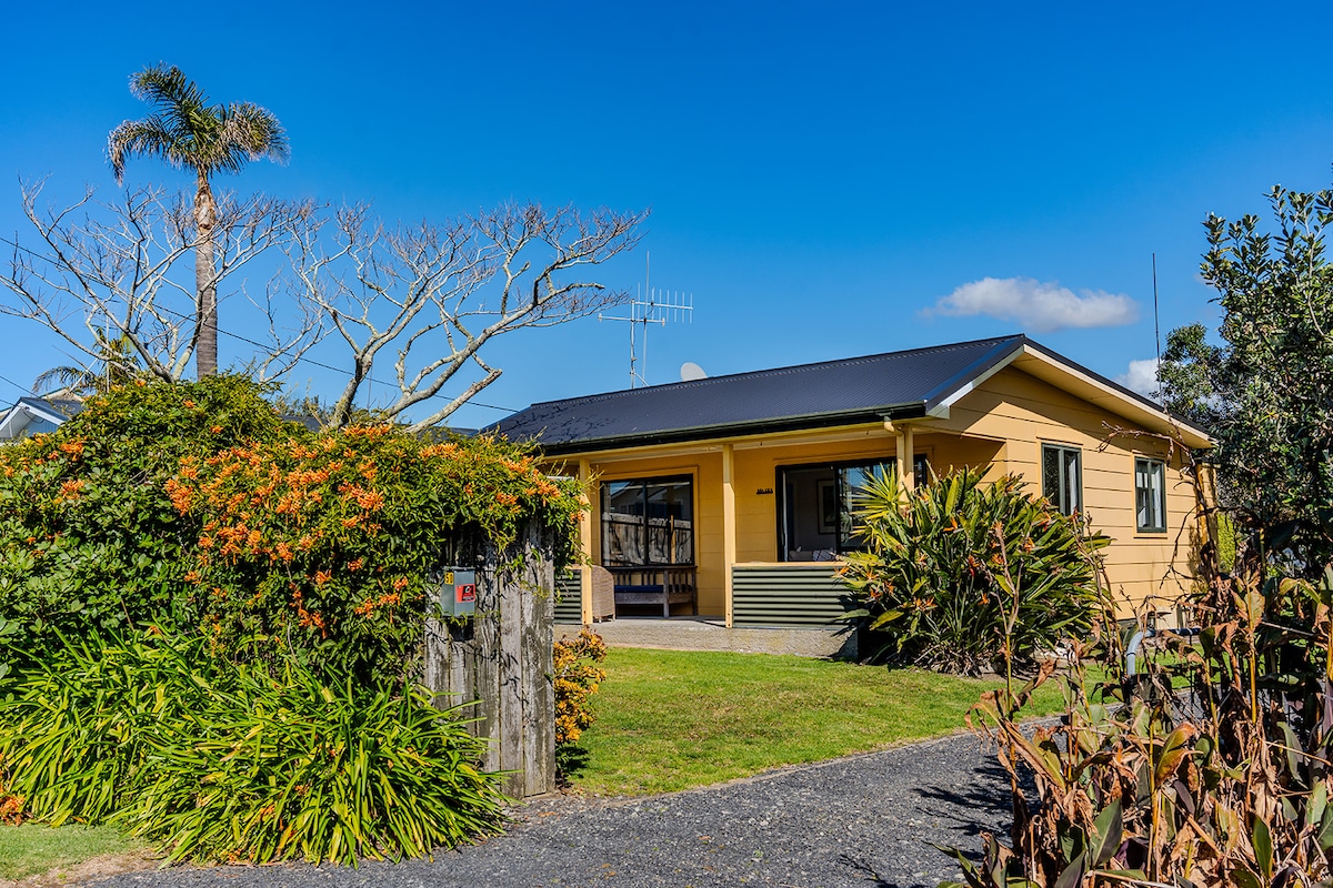 The exterior of a charming beach house is showcased, featuring a warm yellow facade and a well-maintained garden. Lush greenery surrounds the property, with a pathway leading to the entrance. A clear blue sky provides a bright backdrop, enhancing the inviting appearance of the home.