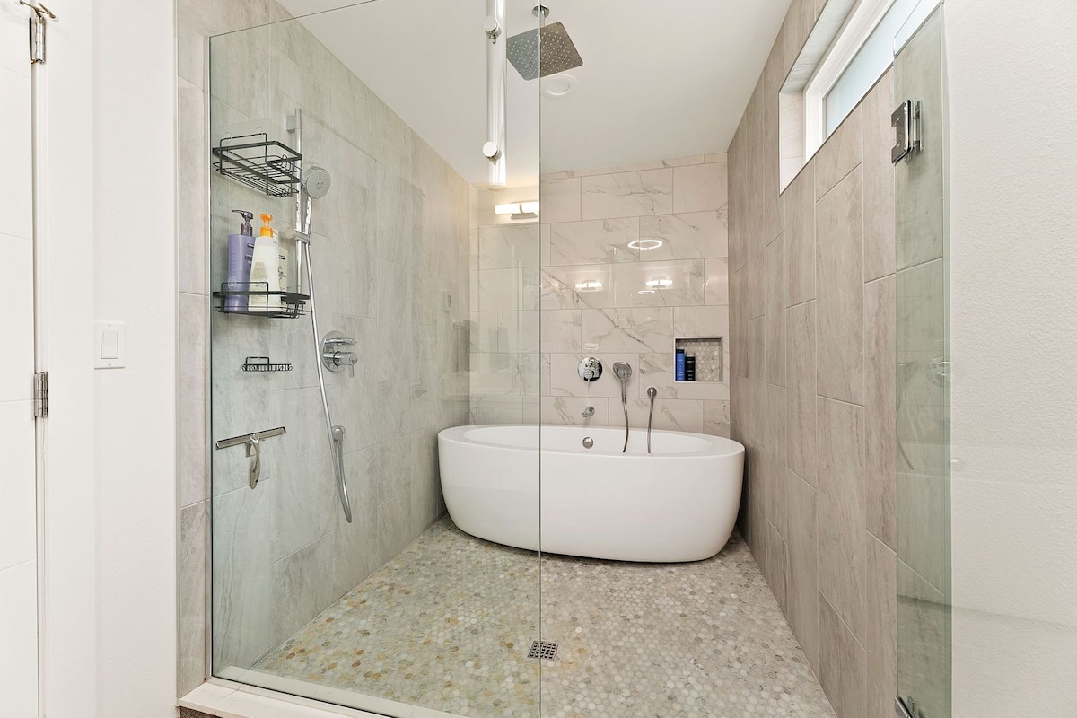 A modern bathroom is presented with a glass-enclosed shower and a freestanding soaking tub. Sleek fixtures and stylish tile work enhance the space. Soft lighting illuminates the room, which features a built-in showerhead and complementary shelves for toiletries.