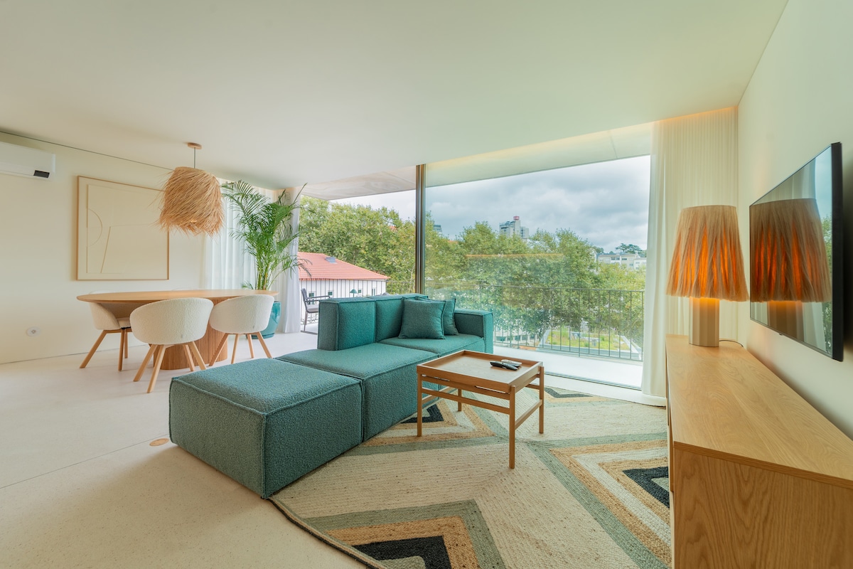 A sunlit living area showcases a spacious teal sofa and a wooden coffee table, positioned on a patterned rug. Nearby, a round dining table with chairs is set against large glass doors that provide views of the surrounding greenery.