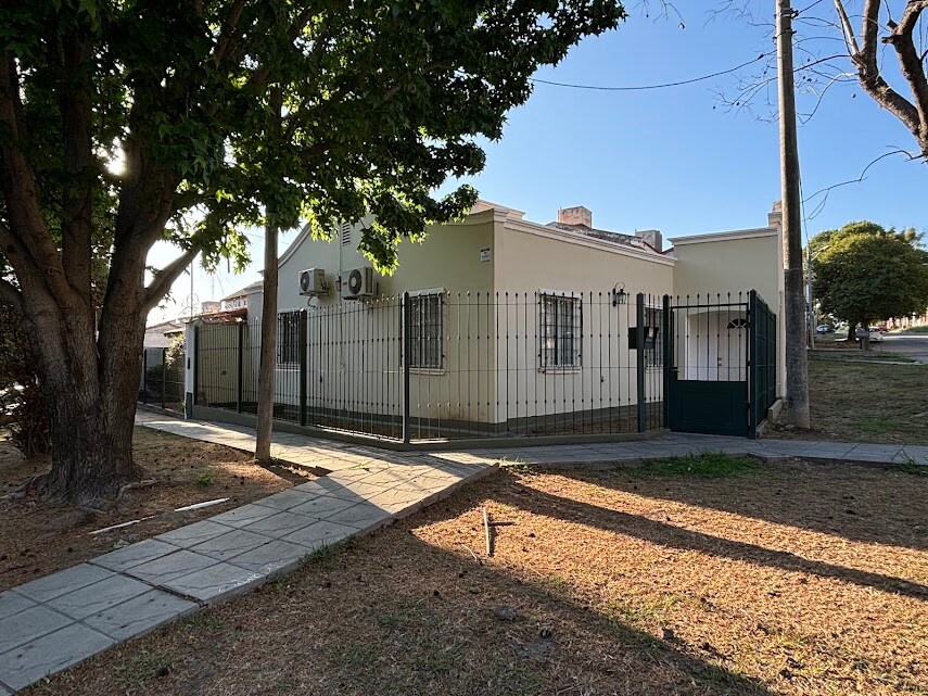 A single-story house is surrounded by a low fence, positioned on a quiet corner lot. Large trees provide shade, while the front walkway leads to the entrance. Air conditioning units are visible on the exterior walls, indicating comfort during warm days.