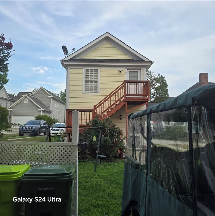 A two-story yellow house features a wooden staircase leading to an upper entrance. A green lawn and outdoor seating are visible in the foreground, accompanied by utility bins and a covered seating area, suggesting a welcoming outdoor space.