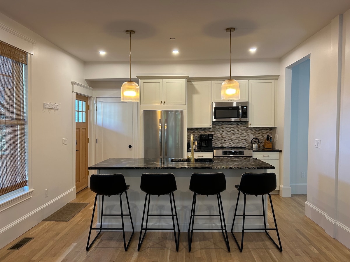 A modern kitchen features white cabinetry and a stone countertop with seating for four. Two pendant lights hang above, providing soft illumination. Stainless steel appliances, including a refrigerator and microwave, are visible. A door to the left offers access to the outside.