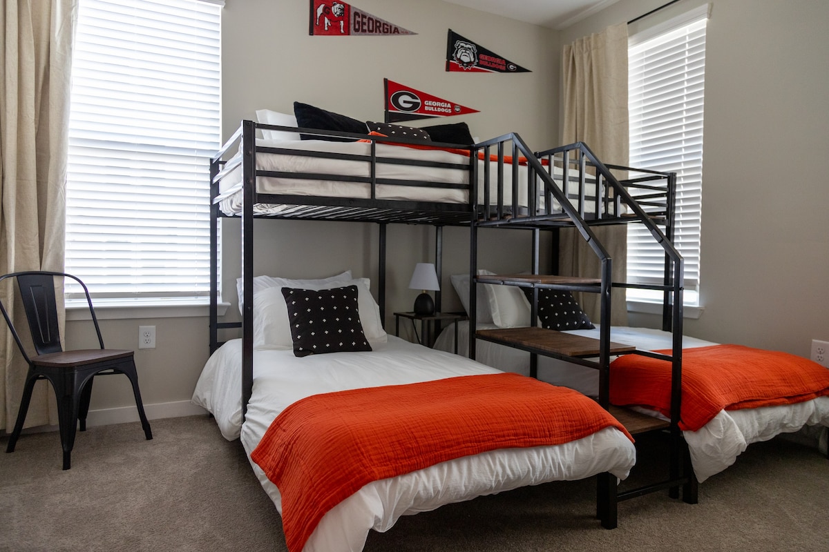 A cozy bedroom features a bunk bed with a spacious bottom bed, complemented by orange blankets. Two windows allow natural light to fill the room, while black curtains frame the space. Decor includes University of Georgia pennants, adding a touch of local spirit.