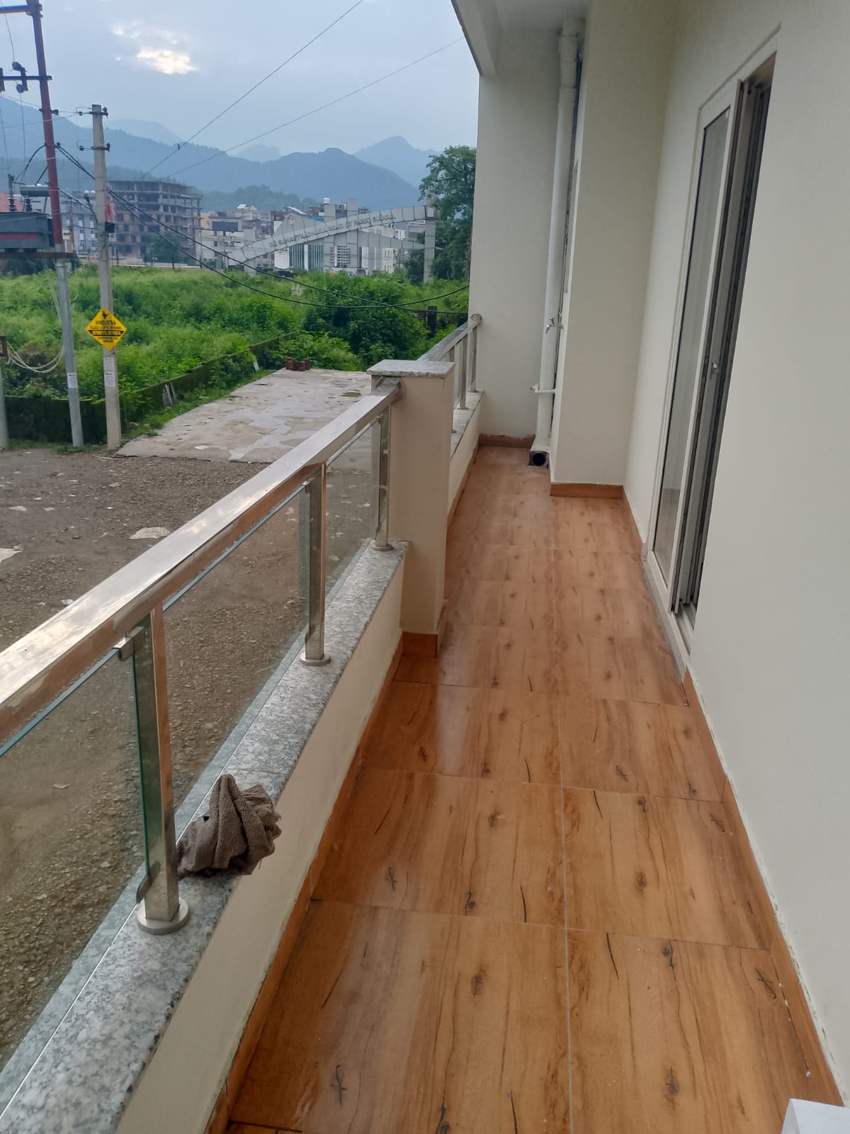 A balcony featuring a wooden floor extends outside, offering panoramic views of nearby greenery and mountains. Glass railings provide safety while allowing unobstructed views. The space is complemented by natural light and a towel casually placed on the railing.